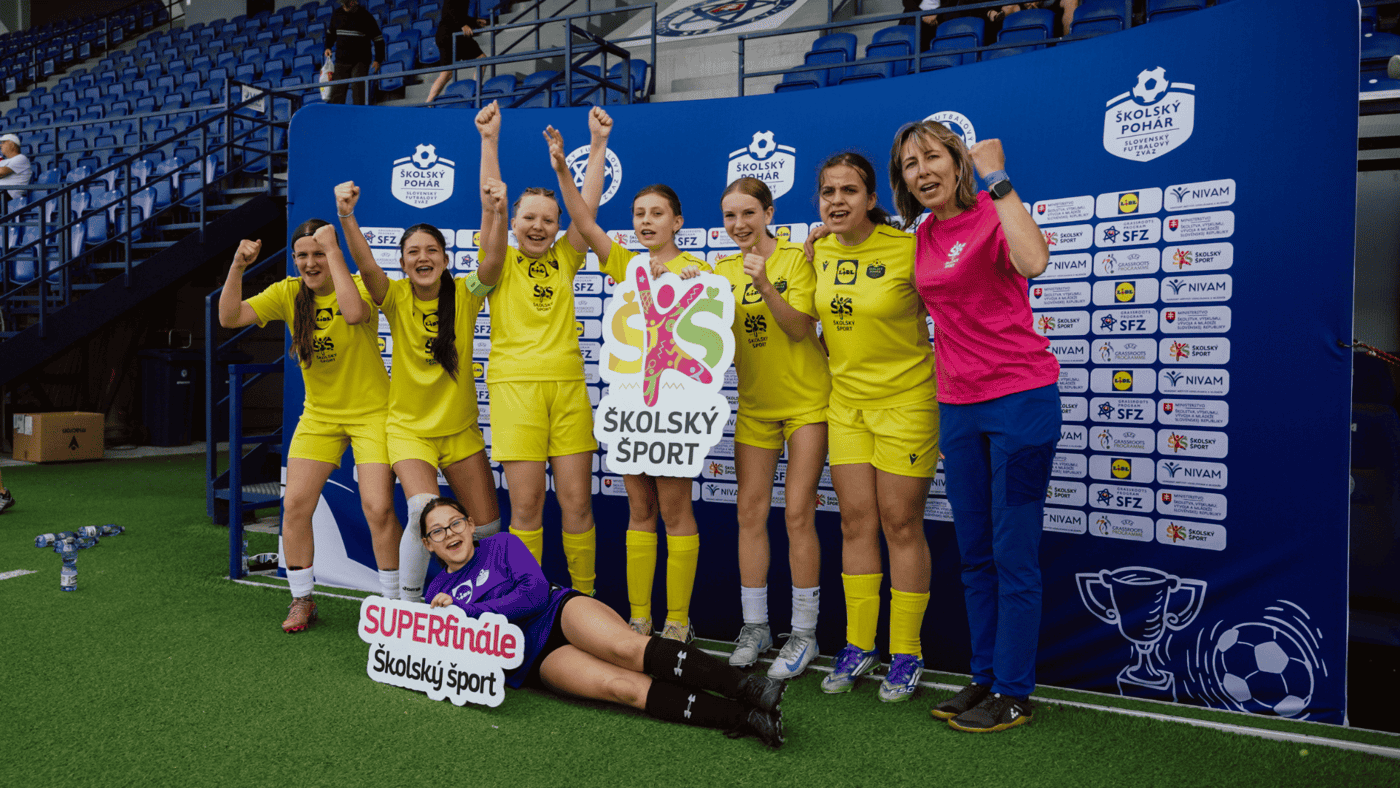 Skupina žiačok v žltých futbalových dresoch stojí pred panelom s logom „Školský pohár“ a „Školský šport“ na štadióne a dvíha ruky na znak víťazstva; jedna hráčka v brankárskom drese sedí vpredu s tabuľkou „SUPERfinále Školský šport“.