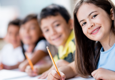 Na fotografii sú štyri deti, ktoré sa pozerajú do kamery a usmievajú. V ruke držia ceruzku a sedia za stolom.