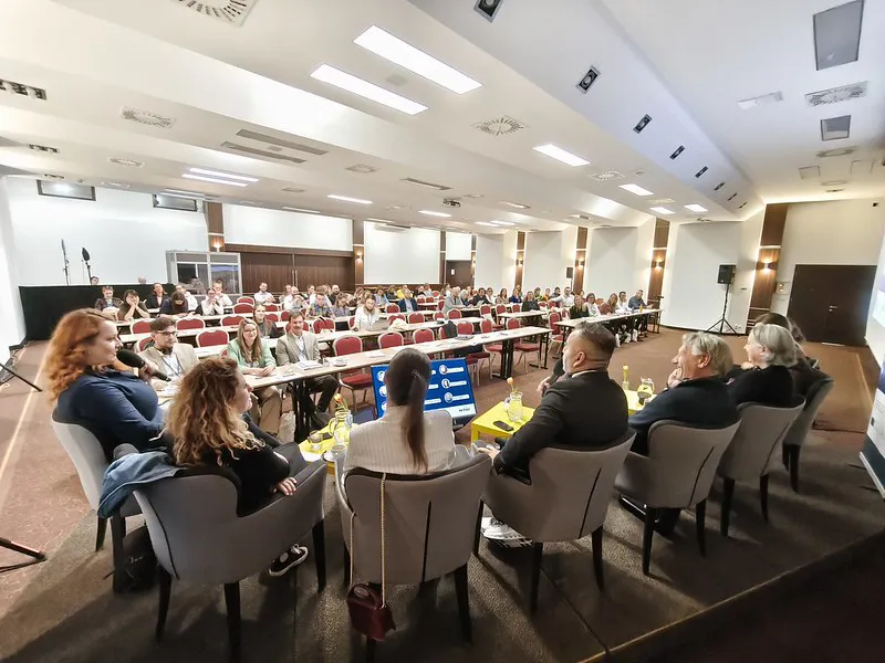 Fotografia z konferencie. Vpredu sedia hostia hovoriaci do mikrofónu, v pozadí obecenstvo.